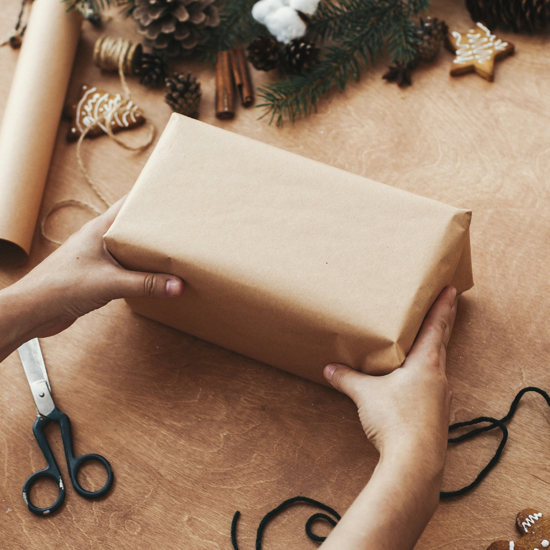 Person wrapping a gift with brown paper on a wooden surface, surrounded by Christmas decorations.