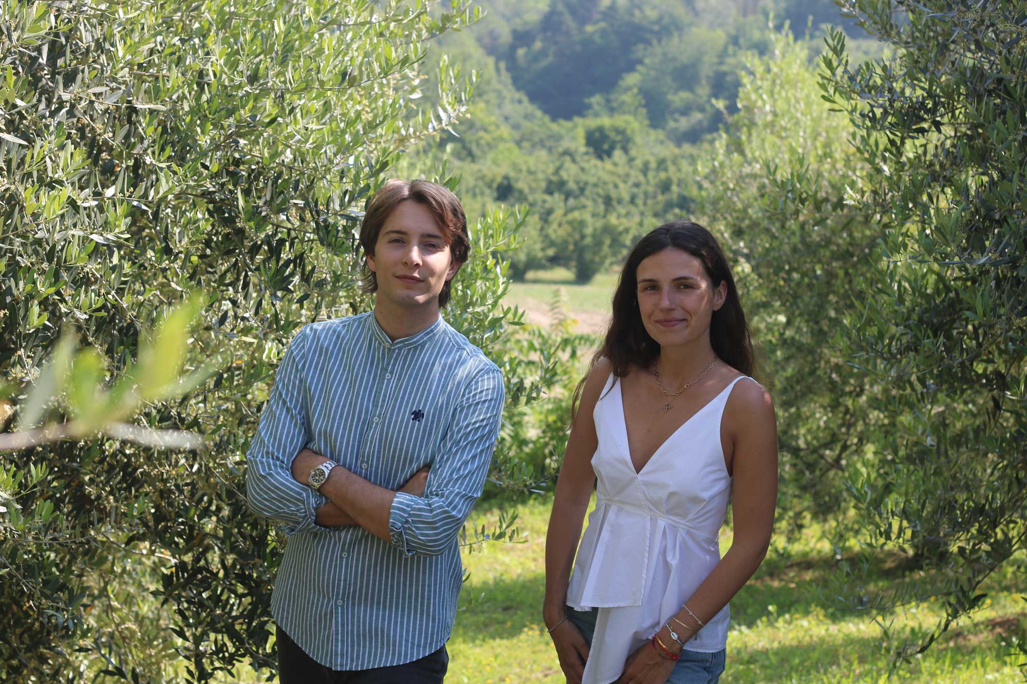 Two people standing in an olive grove with trees and greenery around.